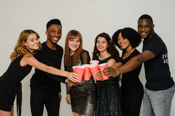 Wall Mural - Waist up of six friends having fun and drinking alcohol during New Year party, isolated on white background. A group of cheerful friends of different nationalities clink glasses with red glasses at a