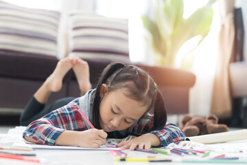 Wall Mural - Happy cute asian little girl drawing using colorful pencils while lying on the floor at home