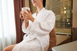 © DragonImages - Close-up of content young woman in white robe sitting on chair and drinking coffee in hotel room