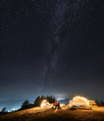 Wall Mural - Two campers looking on evening scenery endless sky covered with stars and Milky Way galaxy from mountain hill where arranged their camp. Summer nightly camping outdoor with shining tents.
