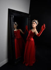 Wall Mural - Full length  portrait of beautiful young asian woman wearing red corset, long opera gloves and ornate crown headdress. Graceful posing against a full length mirror with a dark studio background.