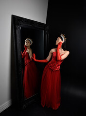 Wall Mural - Full length  portrait of beautiful young asian woman wearing red corset, long opera gloves and ornate crown headdress. Graceful posing against a full length mirror with a dark studio background.