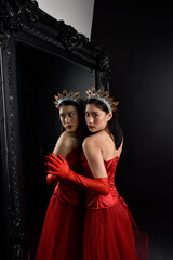 Wall Mural - Full length  portrait of beautiful young asian woman wearing red corset, long opera gloves and ornate crown headdress. Graceful posing against a full length mirror with a dark studio background.