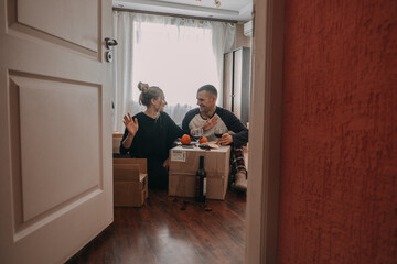 Moving day celebration. Yong couple Move Into New Home. Young happy couple, newlyweds family drinking wine and having fun in room with carton boxes in new home