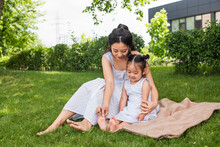 Young Asian Mother Touching Feet Of Toddler Daughter Sitting On Picnic Blanket