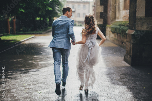 Blurred image of beautiful emotional wedding couple running in rain in european city. Provence wedding. Stylish happy bride and groom walking on background of old church in rainy street.