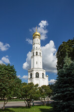 MOSCOW, RUSSIA-AUGUST, 4, 2021: The High White-stone Bell Tower Of Ivan The Great With A Golden Dome Against The Background Of Blue Sky And Green Foliage Of A Tree On A Sunny Summer Day
