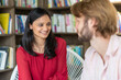 © MandriaPix - Young Indian woman chatting with a Caucasian man, college students flirting in the library, smiles and seductive look at an informal date