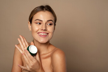 Wall Mural - Cute woman holding a jar of moisturizer in the studio. Skin care and hydration concept. Portrait of beautiful woman applying some cream to her face for skin care