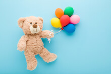 Smiling Brown Teddy Bear Holding Heap Of Colorful Balloons On Light Blue Table Background. Pastel Color. Closeup. Congratulation Concept. Top Down View.