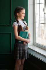 Wall Mural - A cute girl in a school uniform stands by the window with a book in her hands