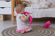 A one-year-old girl in a white and pink openwork dress licks her mother's women's hot pink high-heeled pumps while sitting on the floor in the children's room