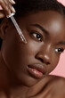 Portrait close up of beautiful black girl dropping serum collagen moisturizer on face. Serious young woman. Concept of face skin care. Isolated on pink background. Studio shoot