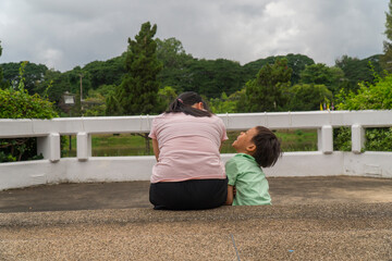 Back view of loving Asian mother hug her kids sitting on bridge, caring black mom embrace child, relaxing looking to black cloud, parent comfort child caressing.