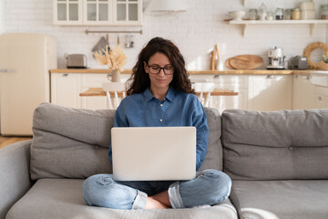 Millennial woman in eyeglasses use laptop computer sitting on couch in living room. Young freelancer worker or student girl shopping online, work remotely or browsing web from home with notebook pc