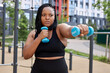 © Roman - Serious and healthy obese african woman exercising with dumbbells, outdoor sport activity concept. Mixed race confident lady in black tracksuit engaged in sport, fighting, concentrated on workout