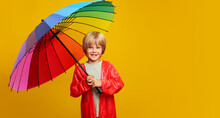 Cheerful Child Boy In Red Raincoat Under A Colored Umbrella On Colored Yellow Background. Copy Space