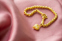 The Gold Bracelet Has A Heart Pendant Placed On A Pink Gold Fabric.