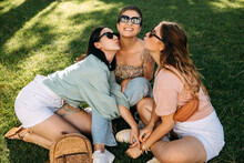 Four Young Women Outdoors, Having Good Time, Sitting On Green Grass, Laughing, Wearing Sunglasses.