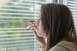 © somemeans - Young woman looks out window through the blinds. Girl with glasses looks at street through blinds.