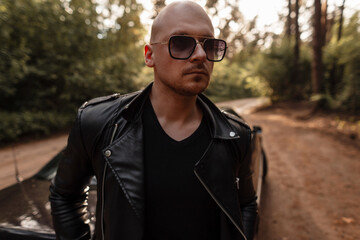 Portrait serious bald man with beard in fashionable black leather clothes in stylish sunglasses on nature at sunset. Attractive american guy rest near modern black car on sandy road in forest.