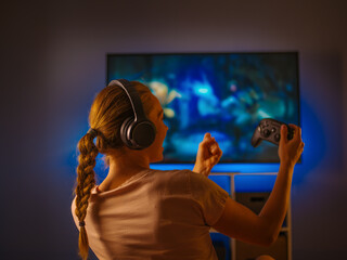 Wall Mural - A young girl plays shooting games with headphones on her head and a joystick in her hand. Shooting from the back. Video games, shooting games, advertising and gaming business.