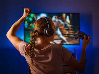 Wall Mural - A blonde girl with headphones and a joystick sits in front of a computer monitor and plays a video game. She reacts emotionally to what is happening on the screen. Gamer, hacker, video games.