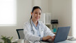 © ChayTee - Young adult asia people or medic staff woman sitting toothy smile arm cross look at camera happy work on desk in clinic office room in telehealth telemedicine smart health care consult online service.