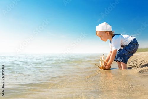 Kid play on the beach on a hot sunny day. Little girl dressed as a sailor stands barefoot on the sandy shore and launches a boat into the sea. Child dreams of travel and adventure.