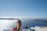 Fototapeta  - Young woman in sun glasses with bouquet of beautiful flowers on terrace of luxury hotel on Santorini island. View of caldera. Aegean sea.