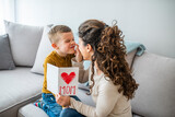 Fototapeta Panele - Happy little boy congratulating smiling mother and giving card with red heart during holiday celebration at home. Cheerful mother hugging son and reading handmade greeting card
