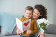 © Dragana Gordic - Cheerful mother hugging son and reading handmade greeting card with heart while resting on bed during holiday celebration mothers day at home.