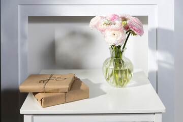 Wall Mural - A bouquet of pink Persian buttercups in a glass vase and two wrapped gifts on a white table in front of a fake white fireplace