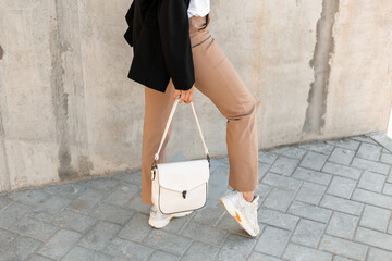 Woman with a fashionable white bag and white stylish sneakers near a gray wall on the street. Women's handbag collection