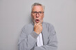 © wayhome.studio  - Studio shot of stunned bearded grey haired elderly man holds chin stands speechless being amazed by something wears spectacles and jacket isolated over grey background looks scared or nervous