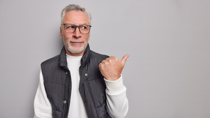 Wall Mural - Serious grey haired aged man points thumb aside shows logo on copy space wears spectacles white jumper and vest poses against grey studio background. Place your advertisement here. Look at this