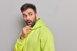 © WHstudio Leushin N - Startled bearded young man holds chin opens mouth from amazement stares speechless in awe cannot believe his eyes wears comfortable green hoodie poses against grey background copy space on right