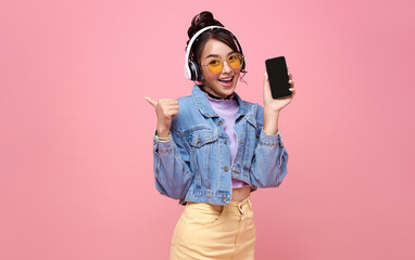 Young Asian teen woman showing smart phone she listening music in headphones isolated on pink background.