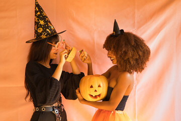 Young Hispanic and Latina women dressed as witches pulling candy out of a pumpkin on a pink background for Halloween.