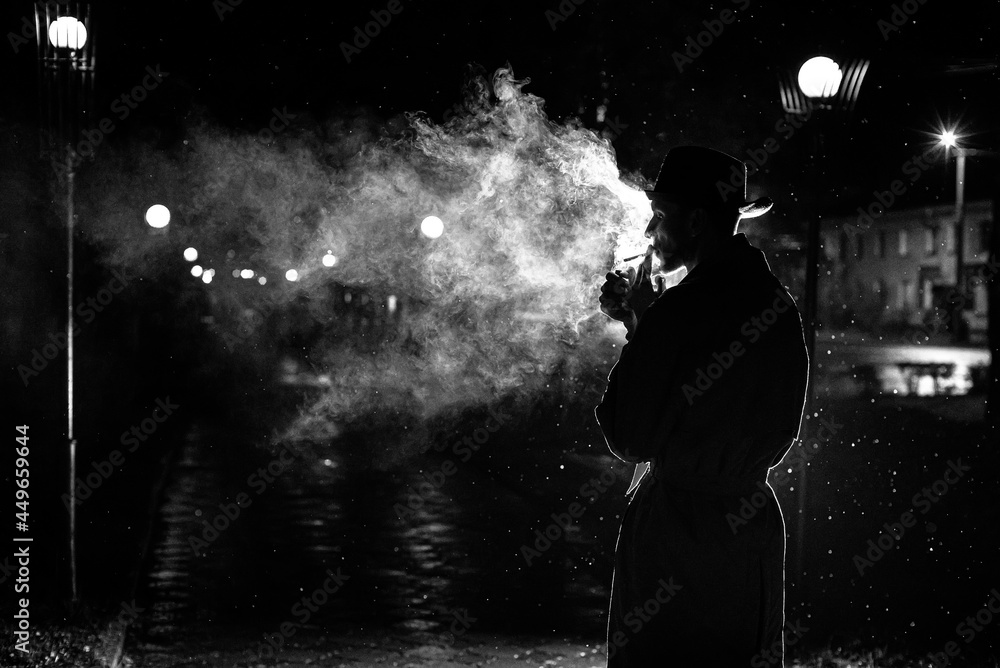 smoke a cigarette in the rain meaning dark silhouette of a man in a hat Smoking a cigarette in the rain on a  night street Stock Photo