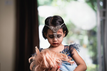 Wall Mural - Little girl in halloween costume holding doll sitting on chair.