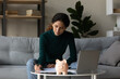Satisfied woman checking finances, calculating domestic bills at home, filling financial documents, writing, sitting at table with pink piggy bank and laptop, sitting on couch, investment concept