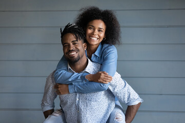 Wall Mural - Portrait of overjoyed millennial biracial woman piggyback hug smiling ethnic husband man. Excited young African American couple family isolated on grey wall background embrace show love and care.