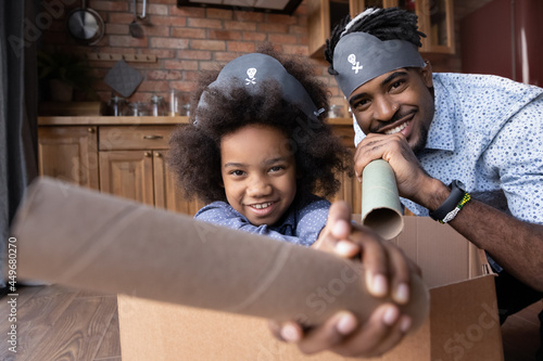 Portrait of overjoyed young African American dad and teen daughter play pirates in ship together at home. Smiling loving ethnic father and small girl child have fun involved in playful game activity.