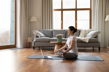 Canvas Print - Calm millennial Indian woman yogi sit on mat meditate with mudra hands have webcam virtual event on computer. Young ethnic female train exercise online on video call on laptop, practice yoga.