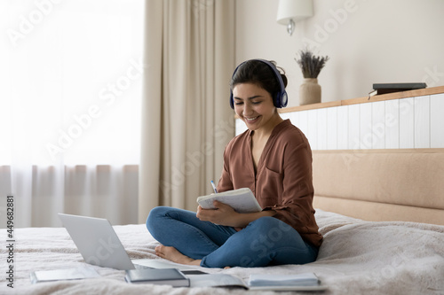 Smiling millennial teen Indian female student in headphones sit on bed at home take note study online on computer. Happy young ethnic woman in earphones busy take distant course or training on laptop.
