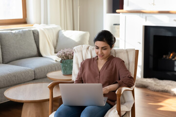 Wall Mural - Young Indian woman sit in chair at home browse wireless internet on modern laptop gadget. Mixed race female relax in armchair use computer work or study distant on device. Technology concept.