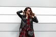 Beautiful young woman in fashion casual red-black youth outfit in stylish sunglasses with leather bag straightens hairstyle near modern wall outdoors. Urban pretty fashionable girl model in city.