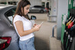 © Aleksandr - Attractive young woman using her phone and leaned on her car at gas station. Woman waiting gas station worker