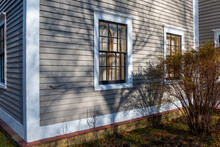 Two Double Hung Windows With Black Wood Frames, Multiple Panes Of Glass, In A Tan Color Wooden Wall. The Wall Has Narrow Clapboard With White Trim. There Are Two Green Bushes In Front Of The Windows.